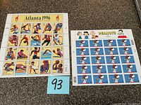 Two sheets of stamps displayed on a brown surface with a lot number card '93'. One sheet has colorful stamps of athletes from the Atlanta 1996 Olympic Games. The other sheet has stamps with blue background featuring Snoopy and Peanuts characters.
