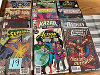 Photo of a collection of 12 comic books arranged on a table including Superman, Batman, Justice League of America, Iron Man, Ka-Zar, Micronauts, and Realm of Kings Nova titles.