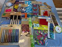 Various craft items including glitter glue tubes, paint brushes, foam shapes, beads, sequins, ribbons, and fabric patches spread on a table.