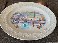 White oval ceramic Christmas platter with a raised decorative border and colored winter village scene in center.