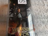 Figurine of Elvis Presley in his Jailhouse Rock outfit within original plastic packaging. Clear shot of figure with arm raised and mouth open, dressed in black leather jacket and striped shirt.