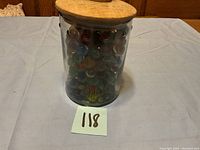 Front view of clear glass jar filled with assorted colorful glass marbles, topped with round wooden lid.