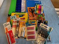 Photo of various school and office supplies arranged on a surface including notebooks, folders, writing instruments, erasers, scissors, and glue.