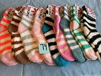 Photo showing 9 pairs of fuzzy slipper socks in a range of colorful stripes placed in a row on a blue surface with tags visible.
