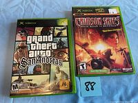 Front view of two Xbox game cases: Grand Theft Auto San Andreas and Crimson Skies High Road to Revenge on white surface.
