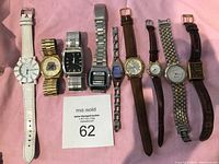 Nine wristwatches positioned horizontally on pink fabric surface showing front face and straps of each watch for identification and condition assessment.