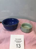 Front view of the two Asian teacups, one blue and one green, on pink cloth with auction tag.