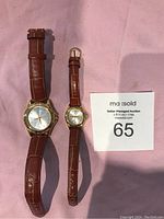 Two Gold Wings branded wristwatches side by side on a pink background with a salesman tag number 65.