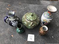 Photo showing five porcelain pieces on a concrete surface: tall white vase with blue floral design, small green vase, green decorative pot with lid, large multicolored floral vase, small brown-gold vase.