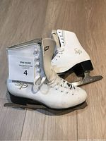 Pair of white figure ice skates with logos SC and SP on sides, laced up, shown on wooden floor.