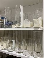 Multiple clear glass cylinder vases on white shelves, various sizes, many with hardened white wax residue inside.