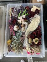 Top-down image of a large clear plastic bin filled with assorted dried florals such as dark red roses, baby's breath, cotton branches, and potpourri sachets, showing the variety and condition of the florals.