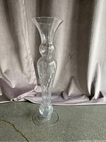 Single large clear glass trumpet vase with flared top and rounded mid-section, approximately 18 inches tall, on floor against curtain.