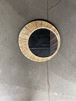 Round mirror with woven or natural fiber circular frame, placed flat on a concrete floor, about 16 inches in diameter.