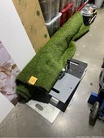 Photo of large rolled green artificial turf rug placed on a black platform on the floor, corners slightly bent.