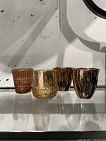 Various votive holders displayed on a shelf showcasing multiple styles including ribbed glass and hammered metallic surfaces in rose gold tone.
