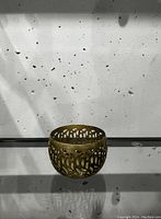 Single gold metal votive holder with openwork cutout design shown on glass shelf with white speckled background.