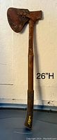 Photo of a 26 inch hatchet/axe with a rusty metal handle and leather blade cover, showing its full length against a white wall background.