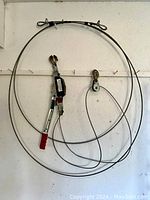 Full view of the heavy duty ratchet winch set hanging on a white wall with the coiled steel cable and hooks visible.