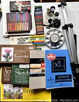 Top view of assorted pastels, sketchbooks, watercolor pads, paint palette and compressor sprayer kit components.