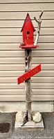 Front view showing the red birdhouse, carved wood post, welcoming sign, pine cone, and natural twig detail.