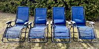 Four blue zero-gravity lounge chairs arranged side-by-side outdoors on grass in front of a hedge.