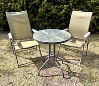 Photo showing two beige sling patio chairs and a round glass-top table on grass with trees in background.