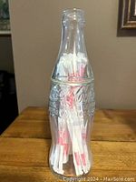 Glass Coca-Cola bottle used as a straw holder with white and red paper-wrapped straws inside, placed on a wooden surface.