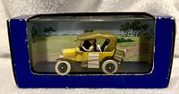 Side view of the yellow Ford Model T toy car featuring Tintin and Snowy inside clear display case with illustrated savanna background.