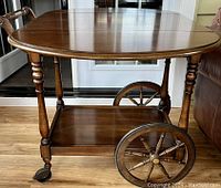 Front view of vintage walnut tea cart showing the top with two drop leaf panels, large spoked wooden wheels on one side, casters on the other, wooden legs and lower shelf.
