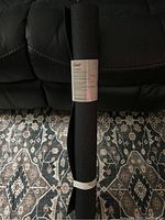 Rolled black rubber mat with label showing brand Master Chef, placed on patterned rug and black leather couch background.