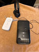 Photo showing TP-Link WiFi booster, Sony headphones, and GE portable tape recorder arranged on a light brown fabric surface.