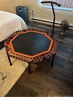 Photo showing the full trampoline with orange cords around the frame and the black handle bar, in a room with wood floor and fan in background.