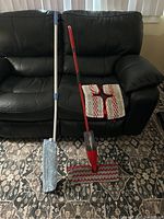 Full view of two floor cleaning tools: a gray and white handled mop with microfiber mop head, and a red handled Vileda spray mop with attached microfiber pad. Also visible are extra microfiber mop pads placed on a black leather couch.