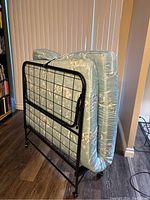 Folded cot and mattress standing upright showing metal frame with grid design and wheels.