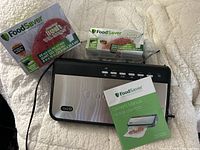 Top view of FoodSaver vacuum sealer with two packages of FoodSaver plastic bags and owner's manual on a textured fabric surface.