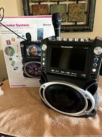 Front view of Karaoke USA GF842 karaoke machine with microphone attached, showing screen, speaker, and control knobs.