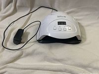 White Sun X7 Plus UV LED nail lamp with attached power adapter on a light-colored fabric background.