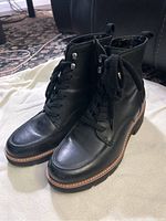 Pair of new black leather lace-up ankle boots displayed on a white cloth with visible stitching and thick sole with brown trim.