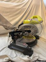 Side view of Ryobi miter saw with blade guard and dust collection bag attached. Clear plastic guard and horizontal handle visible.