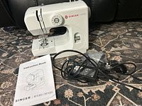 Singer M1000 sewing machine, power adapter and cords, instruction book on patterned carpet.