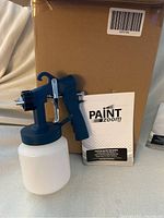 Photo of the Paint Zoom electric mini paint sprayer with attached white container and manual in front of a cardboard box.
