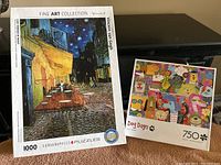Two jigsaw puzzles standing upright: one is a 1000-piece EuroGraphics Fine Art Collection puzzle featuring Vincent van Gogh's 'Café Terrace at Night' painting; the other is a 750-piece Dog Days puzzle with colorful illustrated dogs.