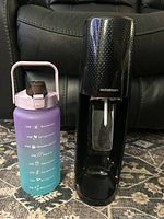 Photo of black SodaStream soda maker and gradient purple-blue water bottle with time-marked hydration schedule placed side by side on floor with black couch background.