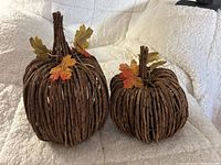Photo of two handwoven wicker pumpkins with attached artificial autumn leaves, placed on a textured cream fabric background.