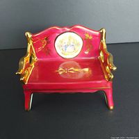 Front angle of red porcelain miniature bench with gold painted accents and a central round Fragonard love story scene on backrest.