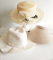 Photo showing three summer hats: a beige woven hat with flowers, a white wide brim hat, and a beige visor arranged on a white surface