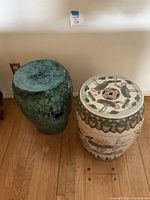 Pair of Asian ceramic garden stool tables photographed side by side on hardwood floor against cream wall, showing details of glaze, patterns, and top surface design.
