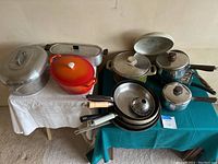 Wide view of assorted cookware and bakeware including pots, pans, and lids on two tables with white and teal tablecloths.