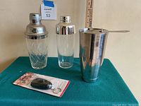 Front view of the three cocktail shakers and the packaged digital breathalyzer on a green table with a ruler for scale